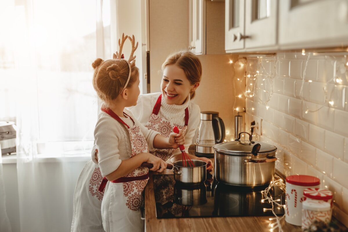 Woman and little girl smiling at each other and cooking at a stove. The little girl is holding a whisk in a sauce pan.