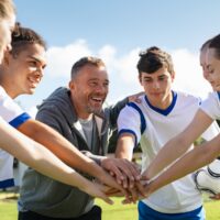 Happy coach and high school soccer team with their hands stacked in football court.